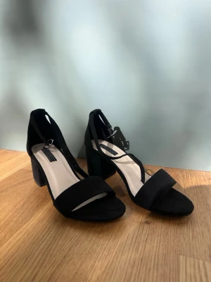 Svarta klackskor NLY Shoes - Stilrena svarta sandaler från NLY Shoes med bred rem över tårna och justerbar rem runt ankeln. Klackarna är blockformade och ger stabilitet. Ovandelen är i mockaimitation och sulan är svart. Helt nya och aldrig använda.