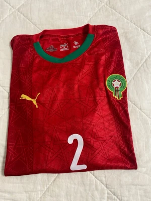 Marocko fotbollströja Puma röd - Snygg röd fotbollströja från Marockos landslag med gröna detaljer vid kragen, gult Puma-märke och landslagets emblem på bröstet. Nummer 2 tryckt på framsidan. Tillverkad i lätt och ventilerande material, perfekt för träning eller match. Köpt från Maroco o är andvänd bara 1 gång. Står 5xl men passar mer som XL