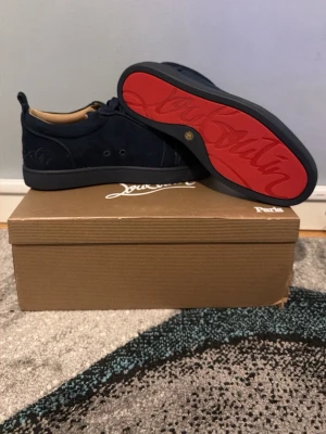 Mörkblå Louboutin sneakers - Snygga mörkblå sneakers från Christian Louboutin med klassisk röd sula och diskret logga på sidan. Skorna har en låg profil och är tillverkade i mocka med svarta snören och beige innerfoder. Perfekta för dig som vill sticka ut med exklusiv stil.