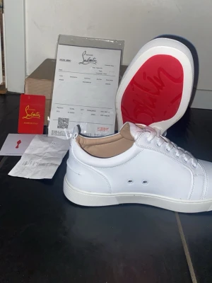 Vita Louboutin sneakers med röd sula - Snygga vita sneakers från Christian Louboutin i läder med den ikoniska röda sulan. Klassisk låg modell med vita skosnören och diskreta ventilationshål på sidan. Insidan är beige och designen är clean och stilren.