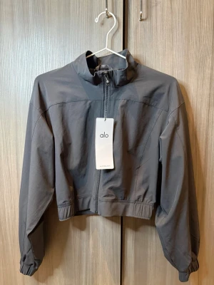 Grå cropped jacka från Alo - Snygg grå cropped jacka från Alo med hög krage och dragkedja framtill. Jackan har resår i midjan och vid ärmsluten för en sportig look. Tillverkad i ett lätt material som passar perfekt till streetwear eller träning.
