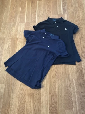 2 Ralph Lauren pikétröjor, M - Två stilrena pikétröjor från Ralph Lauren i storlek M. En är svart och en är mörkblå, båda med klassisk krage, knappar och vit broderad logga på bröstet. Tillverkade i 100% bomull och har slim passform. Mer åt det dam hålet. Hämtas i Värnamo elller skickas med köparens frakt….
