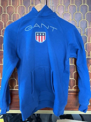 Blå hoodie från GANT med logga - Snygg blå hoodie från GANT med broderad logga och amerikanskt inspirerat märke på bröstet. Klassisk känguruficka framtill och huva. Perfekt för en avslappnad och sportig stil.