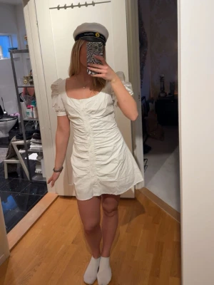 Vit Studentklänning - Snygg vit kort klänning med puffärmar och rynkade detaljer framtill. Klänningen har fyrkantig ringning och är figurnära upptill med en lätt utsvängd kjol. Perfekt för student eller sommarens tillfällen. Aldrig använd från meet me there i storlek S/M