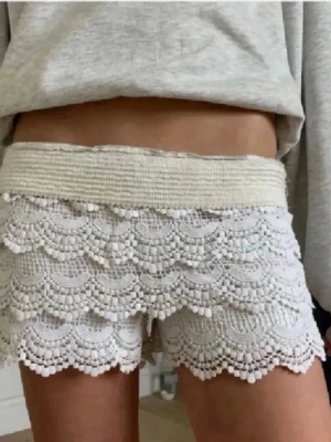 Vita virkade shorts med spetsdetaljer - Säljer ett par vita virkade shorts med bred resår i midjan och vackra spetsvolanger. Shortsen har en feminin look med scallop-kant och små bollar längs kanterna. Perfekta för varma dagar och ger en bohemisk vibe.