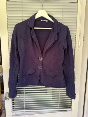 Mörklila kavaj från Gina Tricot - Stilren marinblå kavaj från Gina Tricot med två knappar framtill och två dekorativa fickor. Kavajen har en klassisk krage och är tillverkad i ett mjukt tyg som ger en avslappnad look. Perfekt för dig som vill ha en enkel men snygg kavaj till garderoben. Är blekare en ny i färgen, Står L men är mer som M kanske tillumed S.