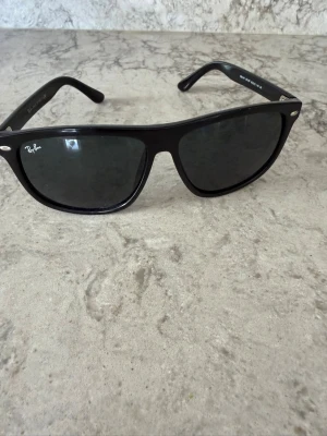 Svarta Ray-Ban Wayfarer solglasögon - Klassiska svarta Wayfarer solglasögon från Ray-Ban med ikonisk fyrkantig form och mörka glas. Snygg och tidlös design med Ray-Ban-logga på linsen och robust båge i plast. Perfekta för att lyfta din stil under soliga dagar.