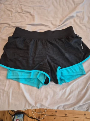 Svarta och turkosa löparshorts - Snygga löparshorts i svart med turkosa detaljer och innershorts. Shortsen har bred resår i midjan och är tillverkade i ett lätt och snabbtorkande material, perfekt för träning och löpning. Sportig design med skön passform.