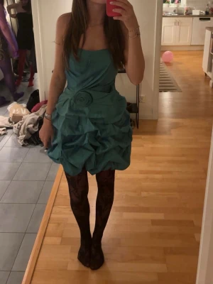Turkos axelbandslös kortklänning - Snygg turkos axelbandslös kortklänning med volangdetaljer och en dekorativ ros i midjan. Klänningen har en ballongliknande kjol och är tillverkad i ett glansigt material som ger en festlig känsla. Använd en gång på en fest med tema the capitol! (Köpt för 900 kr) Har även sytt in under armen inget som syns men värt att nämna. Färgen är blå/grön 