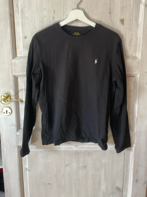 Svart långärmad t-shirt från Polo Ralph Lauren - Svart långärmad t-shirt från Polo Ralph Lauren med klassisk rund hals och den ikoniska lilla loggan broderad på bröstet. Tillverkad i mjuk bomull som känns skön mot huden. Perfekt för en stilren och avslappnad look.