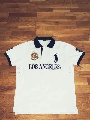 Vit pikétröja Polo Ralph Lauren - Vit pikétröja från Polo Ralph Lauren med svart krage och detaljer. Stor Los Angeles-text framtill, broderad logga och emblem på bröstet samt nummer på ärmen. Klassisk passform och kortärmad design, perfekt till en avslappnad men snygg look.