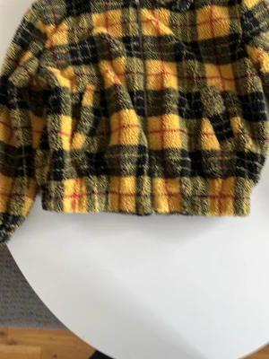 Gulrutigt teddyjacka från New Look - Teddyjacka i mjukt material från New Look med gult, svart och rött rutmönster. Jackan har dragkedja framtill, två fickor och svart slätt innerfoder. Perfekt för dig som vill sticka ut med en färgglad och mysig jacka.