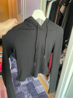 Svart ribbad hoodie med huva - Snygg svart hoodie i ribbat material med långa ärmar och dragsko i huvan. Modellen är croppad och har en tight passform som ger en modern look. Perfekt för dig som gillar stilrena och bekväma plagg. Materialet känns mjukt och följsamt.