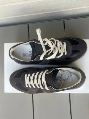 Maison margiela gats - Snygga svarta sneakers med grå mockadetaljer och vita snören. Skorna har en klassisk låg siluett och en brun gummisula. Perfekta för dig som gillar stilrena och tidlösa sneakers med en touch av retro. Materialmix av läder och mocka.