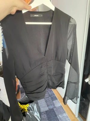 Svart meshblus från Bik Bok - Snygg svart blus från Bik Bok med transparenta meshärmar och v-ringning. Blusen har rynkade detaljer framtill och är tillverkad i ett lätt, tunt material som ger en luftig känsla. 