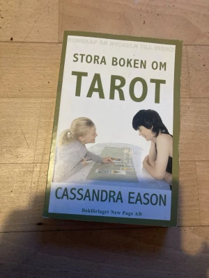 Stora boken om Tarot - Utförlig bok om tarotkort av Cassandra Eason. Omslaget är grönt och vitt med bild på två personer vid ett bord med tarotkort. Perfekt för dig som vill lära dig mer om tarot, tolkningstekniker och utveckla din intuition. Inbunden bok med informativ text och illustrationer.