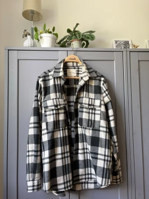 Rutig overshirt från A Day's March - Snygg overshirt i svart och vit rutigt mönster från A Day's March. Skjortan har två bröstfickor med lock, klassisk krage och är tillverkad i ett mjukt, tjockt material som passar perfekt som lager. Perfekt för en avslappnad och trendig look.