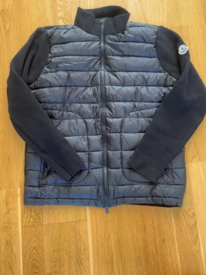 Svart cardiganjacka från Moncler - Snygg svart cardiganjacka från Moncler med quiltad framsida och stickade ärmar. Jackan har dragkedja framtill, hög krage och Moncler-logga på ärmen. Perfekt för lager-på-lager och streetstyle. Säljer eftersom den ej passar mig. Aldrig använd. Kom med bud/frågor!!