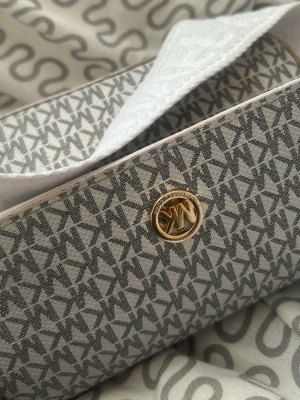 Grå axelväska från Michael Kors - Snygg axelväska från Michael Kors i grått med MK-logga över hela väskan och guldfärgad detalj framtill. Väskan har en bred, vit axelrem med diskret text och är tillverkad i tåligt syntetmaterial. Perfekt för dig som gillar stilrena accessoarer.