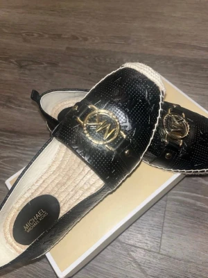 Svarta ballerinaskor Michael Kors - Snygga svarta ballerinaskor från Michael Kors med guldfärgad MK-logga framtill. Skorna har präglat mönster i lädret och naturfärgad flätad sula. Använt max 3ggr.