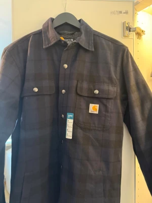 Svart-rutig overshirt från Carhartt - Overshirt från Carhartt i svart och mörkgrått rutmönster. Modellen har relaxed fit, två bröstfickor med lock och tryckknappar samt mjukt teddyfoder på insidan. Klassisk krage och Carhartt-logga på ena fickan. Perfekt för lager-på-lager-stil.