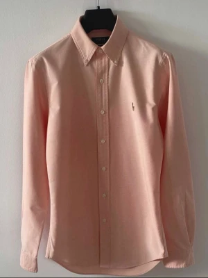 Ljusrosa skjorta från Polo Ralph Lauren - Klassisk ljusrosa skjorta från Polo Ralph Lauren med button-down krage och broderad logga på bröstet. Skjortan har långa ärmar och knäppning framtill. Perfekt för en stilren look.