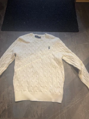 Polo Ralph lauren stickad tröja  - Kabelstickad vit tröja från Polo Ralph Lauren med rund halsringning och den klassiska broderade loggan på bröstet. Tröjan har långa ärmar och ribbade muddar vid ärmslut och nederkant. Perfekt för dig som gillar stilrena och tidlösa plagg.