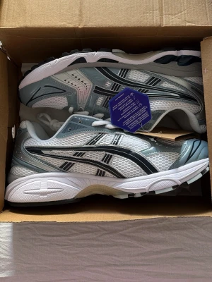 Asics GEL sneakers i vitt och blått - Snygga Asics GEL sneakers med vit mesh och ljusblå detaljer. Skorna har klassisk snörning, silvriga inslag och en sportig vibe. Ovandelen är i ventilerande mesh och syntetmaterial, perfekt för en aktiv livsstil. Sulan är vit med svarta och blå accenter.