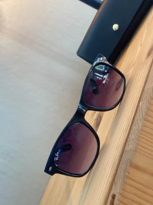 Svarta Ray-Ban solglasögon Wayfarer - Snygga svarta Ray-Ban Wayfarer solglasögon med klassisk fyrkantig form och mörklila tonade glas. Perfekta för att lyfta din stil med en ikonisk accessoar. Ray-Ban-loggan syns tydligt på ena glaset.