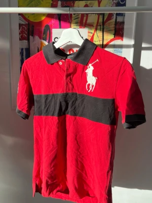 Röd pikétröja från Polo Ralph Lauren - Snygg röd pikétröja från Polo Ralph Lauren med svart krage och bred svart rand över bröstet. Stor vit broderad logga på bröstet och klassisk pikéknäppning. Perfekt för en sportig och stilren look. Storlek L i junior vilket motsvarar S i senior. 