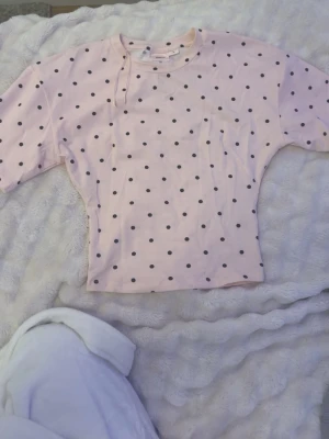 Ljusrosa prickig topp - Söt ljusrosa topp med svarta prickar och rund halsringning. Toppen har korta ärmar och en rak passform, perfekt för en avslappnad och trendig look.