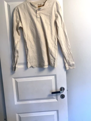 Beige långärmad topp Kappahl  - En stilren beige långärmad topp från Kappahl i mjuk bomull. Modellen har en enkel knäppning vid halsen och rak passform. Perfekt att matcha med jeans eller kjol för en avslappnad look.