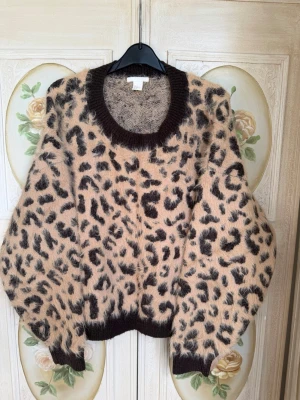 Leopardmönstrad stickad tröja - Supermjuk stickad tröja med leopardmönster i beige och brunt. Tröjan har breda muddar i mörkbrunt vid hals, ärmslut och nederkant. Oversized passform och fluffigt material som ger en cool och trendig look.