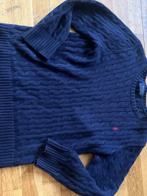 Mörkblå kabelstickad tröja Polo Ralph Lauren - Kabelstickad mörkblå tröja från Polo Ralph Lauren med rund halsringning och ribbade muddar. Klassisk röd broderad logga på bröstet. Perfekt för lager-på-lager och snygg till jeans eller kjol.