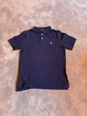 Marinblå pikétröja från Ralph Lauren - Klassisk marinblå pikétröja från Ralph Lauren med vit broderad logga på bröstet. Tröjan har korta ärmar, krage och knäppning med två knappar framtill. Perfekt för en stilren och avslappnad look. Ny pris 800kr