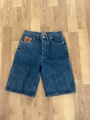 Blå denimshorts från Corteiz  - Snygga blå denimshorts från Corteiz med klassisk femficksdesign och en brun läderpatch med logga på fickan. Shortsen har raka ben och tydliga sömmar framtill för en cool look. Perfekta för varma dagar och enkel att matcha med t-shirt eller hoodie.