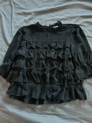 Svart volangblus från Bubbledom - Supersnygg svart blus från Bubbledom med flera lager volanger framtill och volangkrage. Blusen har lång ärm och är tillverkad i ett lätt, flowy tyg som ger en cool och trendig look. Perfekt att styla med jeans eller kjol för en chic outfit.