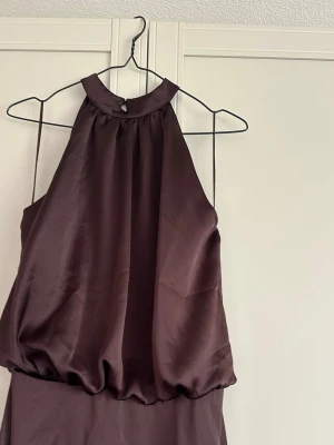 Långklänning - Elegant långklänning i plommonlila satin med halterneck och knäppning i nacken. Klänningen har en mjukt draperad överdel och en slät kjol, perfekt för speciella tillfällen. Sidenliknande material ger en lyxig känsla. Helt ny med lapp. 