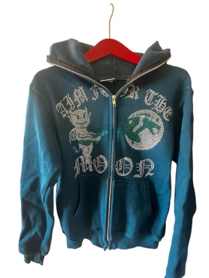 Blå hoodie med strasstryck och dragkedja - Snygg blå hoodie med dragkedja och huva. Framsidan har ett stort strasstryck med texten 'AIM FOR THE MOON' och motiv av en figur och måne. Hoodien har två fickor framtill och ribbade muddar. Perfekt för en cool och avslappnad stil.