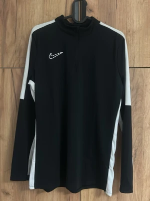 Svart Nike Dri-FIT träningströja - Svart långärmad träningströja från Nike med vita detaljer längs ärmarna. Tröjan har halv dragkedja framtill och är tillverkad i Dri-FIT-material som transporterar bort fukt. Perfekt för träning eller uppvärmning. 