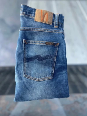 Nudie jeans grim trim  - Snygga blå jeans från Nudie Jeans med klassisk femficksdesign och kontrastsömmar. Jeansen har en ljus tvätt och ikonisk vågdetalj på bakfickorna samt läderpatch med logga i midjan. Perfekta för en avslappnad och trendig look.