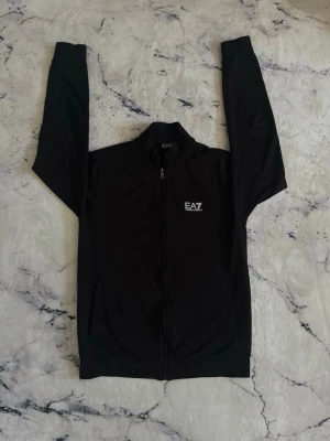 Svart ziptröja från EA7 Emporio Armani - Svart ziptröja från EA7 Emporio Armani med logga på bröstet och diskreta ränder i nacken. Tröjan har hög krage, hel dragkedja och ribbade muddar. Perfekt för en sportig och stilren look.