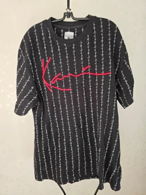 Svart t-shirt från Karl Kani - Svart t-shirt från Karl Kani med vita vertikala ränder och stor röd logga broderad framtill. Oversized passform och rund halsringning. Tillverkad i mjuk bomull som känns skön mot huden.