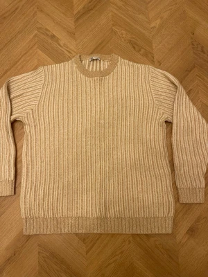 Beige ribbstickad tröja - Snygg beige ribbstickad tröja med rund hals och långa ärmar. Tröjan har ett diskret randigt mönster i olika nyanser av beige och breda ribbade muddar vid ärmslut och nederkant. Perfekt för lager på lager och enkel att matcha.