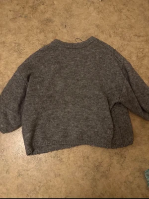 Grå stickad tröja från Gina Tricot - Mysig grå stickad tröja från Gina Tricot med rund halsringning och avslappnad passform. Tröjan har långa ärmar och ribbstickade muddar vid hals, ärmslut och nederkant. Perfekt för lager-på-lager under kyliga dagar.