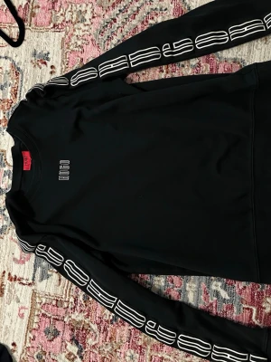 Svart sweatshirt från Hugo Boss - Cool svart sweatshirt från Hugo Boss med vit logga på bröstet och grafiska detaljer längs ärmarna. Tröjan har rund halsringning och ribbade muddar. Perfekt för en avslappnad och trendig stil.