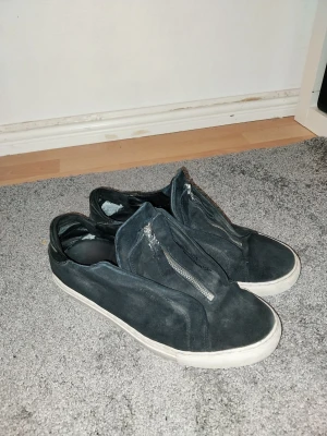 Axel Arigatos dragkedja - Svarta sneakers i mocka från Axel Arigato med vit sula och cool dragkedja på ovansidan istället för klassiska snören. Snygg minimalistisk design   Skorna är använda i vanligt bruk dom är ganska slitna men fungerar att använda som ett par vardagliga skor perfekt för dig som vill ha ett par arigatos men inte kan betala högsta peng då dom fortfarande ser bra ut från längre håll.      Dom har sett ut såhär i nån månad och går nog inte att bli mycke mer förstörda. Storlek 43 passar dig som har 44 bra