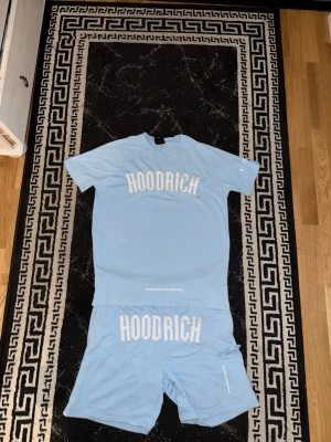 Ljusblått set från Hoodrich - Säljer ett ljusblått set från Hoodrich med matchande t-shirt och shorts. Båda plaggen har tydlig vit logotyp framtill. T-shirten har klassisk passform och rund hals, shortsen har resår i midjan och fickor på sidorna. Perfekt för en avslappnad stil.