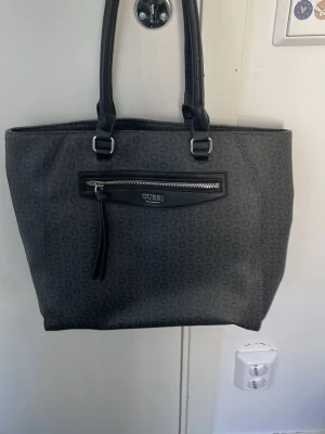Svart axelväska med logomönster Guess - Stilren svart handväska från Guess med logomönster över hela väskan. Väskan har dubbla handtag, en praktisk ytterficka med dragkedja och Guess-logga.  Tillverkad i konstläder med textilfoder. Med en matchande plånbok med många plocka fack. Aldrig använd som helt ny. Har försökt kolla på hemsidan nypriset men hittade inte men skulle gissa runt 1500. Pris kan diskuteras.
