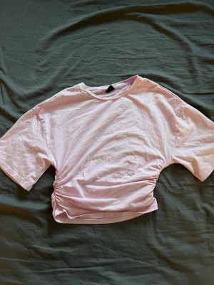 Ljusrosa tight t-shirt Gina Tricot - Trendig ljusrosa t-shirt från Gina Tricot med tight midja passform och snygg  i sidan. T-shirten har korta ärmar och är gjord i mjuk bomull, perfekt för en avslappnad och cool look.
